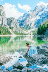 Woman in long blue dress stay on stone near  braies lake in south tyrol, italy. Reflection in the water.