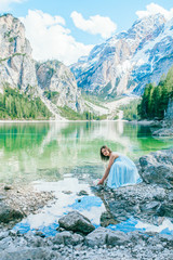 Woman in long blue dress stay on stone near  braies lake in south tyrol, italy. Reflection in the water.