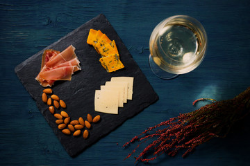 Snacks on a slate tray, with glass of white wine at dark blue background. Cheeses, nuts, prosciutto. Toned image, author processing