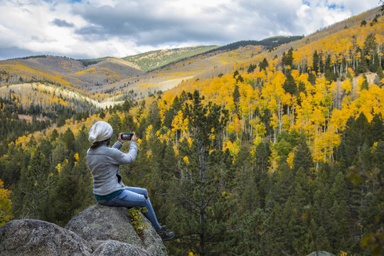 woman taking picture with cell phone of beautiful mountains covered in fall colors