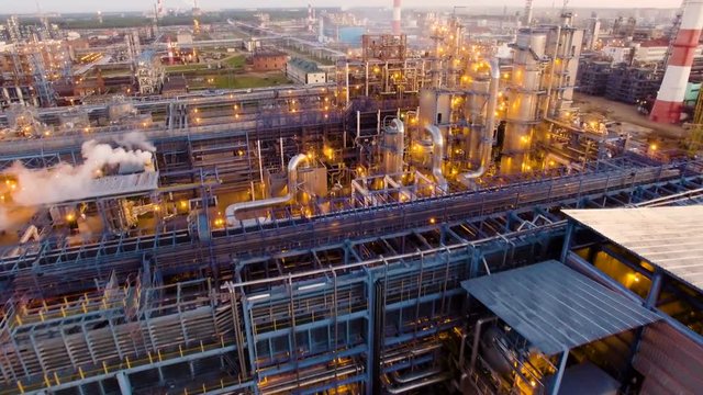 A huge oil refinery with metal structures, pipes and distillation of the complex with burning lights at dusk. Aerial view