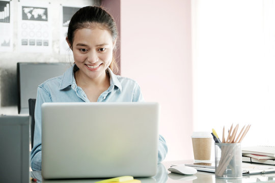 Young asian businesswoman working with laptop computer at office table with smiling face, positive emotion, casual office life concept