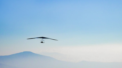 Hang glider soaring the thermal updrafts suspended on a harness below the wing, misty mountain peaks and blue sky