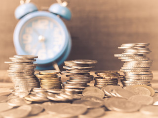Closeup coins stacked on each other in different positions with alarm clock and wooden background.. Money saving concept.