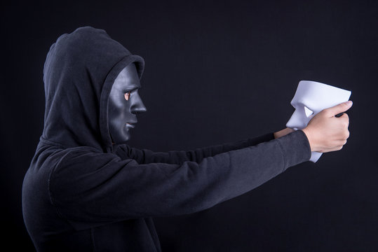 Mystery man wearing black mask holding and looking at white mask. Anonymous social masking or halloween concept.