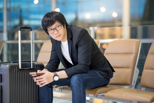Young Asian well-dressed businessman holding passport sitting on bench near his suitcase luggage while waiting for connecting flight in airport terminal. man in business travel concept