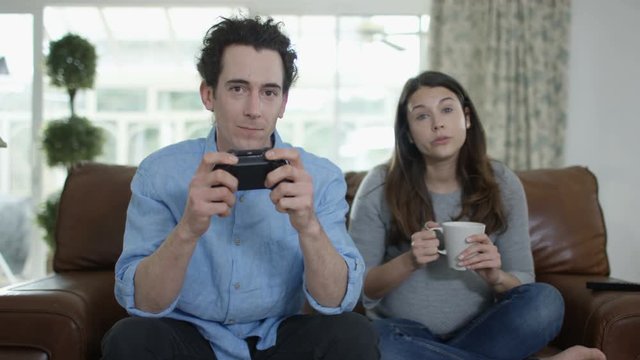  Competitive man playing video games at home, with bored pregnant wife