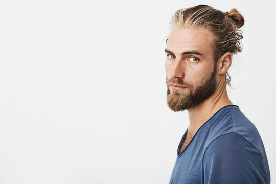 Close up of manly handsome guy with fashionable hairstyle and beard looking in camera, holding head in three quarters with serious expression.
