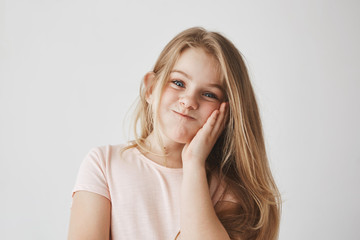 Funny little girl with beautiful long blonde hair looking in camera, squeezing cheek with hand, making funny face expressions, having fun..