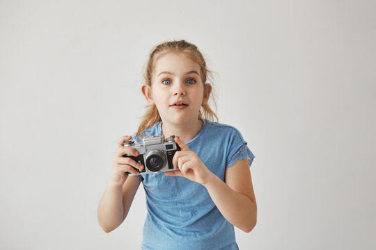 Portrait of good-looking blond girl in blue t-shirt holding camera in hands with concentrated expression, going to take a picture of cute cat on street.