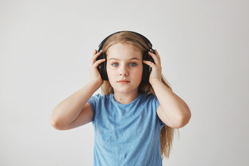 Portrait of beautiful blonde little girl with long hair and blue eyes wearing big headphones, holding it with hands, listening to music with relaxed expression.