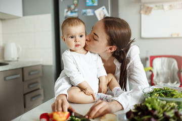 Portrait of happy beautiful mother kissing her lovely baby in cheek in dinner room. Baby sitting on table with surprised expression.