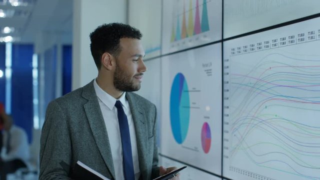 Portrait smiling businessman standing next to video wall with graphs & data