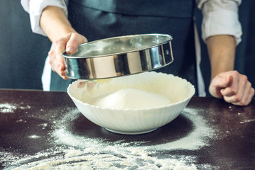 The chef in black apron sifts the flour through a sieve to prepare the dough for pizza