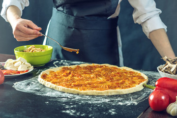 The chef in black apron makes pizza with his hands putting the ingredients for the pizza on the table.