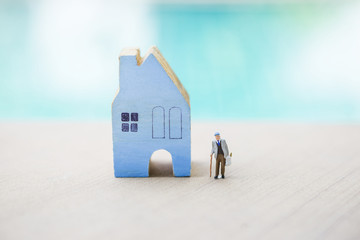 Miniature old man carry grocery bag with miniature wooden house over blurred blue background 