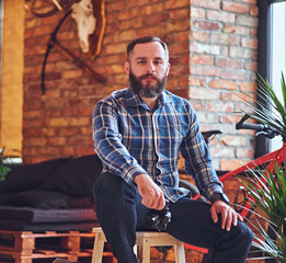 A bearded hipster male dressed in a fleece shirt sits on a wooden chair.