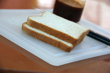 The bread on the white chopping board and out focus knife and jam on the wooden table. Preparation of simple breakfast on the morning.