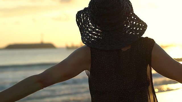 Woman in straw hat standing next to the sea during sunset, steadycam shot

