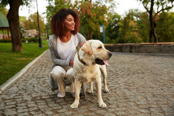Young smiling lady in casual clothes sitting and hugging dog in park