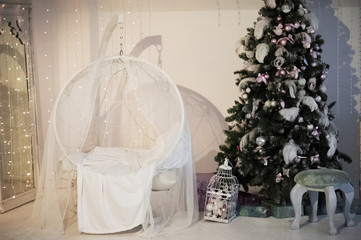 The room is decorated for Christmas and New Year in white style. Christmas tree decorated with feathers, balls, white armchair, Bengal lights, Christmas garland