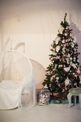 The room is decorated for Christmas and New Year in white style. Christmas tree decorated with feathers, balls, white armchair, Bengal lights, Christmas garland