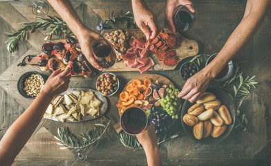 Flat-lay of friends eating and drinking together. Top view of people having party, gathering, celebrating at wooden rustic table set with various wine snacks and fingerfoods. Hands holding glasses - obrazy, fototapety, plakaty