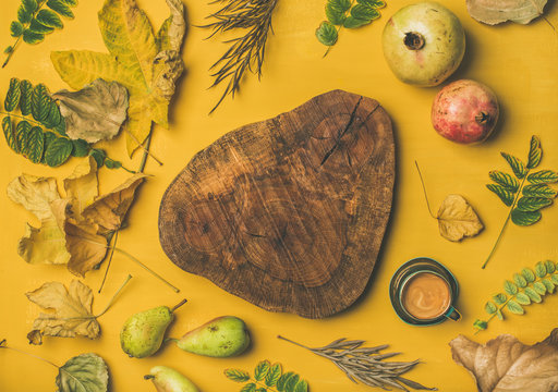 Autumn morning coffee concept. Flat-lay of cup of espresso and wooden board in center over mustard yellow background with dried fallen leaves, pomegranates and pears around, top view, copy space