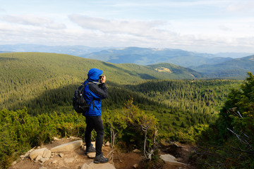 Young professional traveller man with dslr camera shooting outdoor fantastic mountain landscape. Hiker stands on a rock at the high summit and photographing