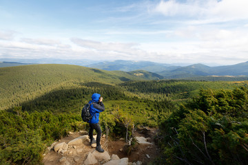 Naklejka premium Young professional traveller man with dslr camera shooting outdoor fantastic mountain landscape. Hiker stands on a rock at the high summit and photographing