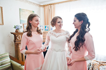 Fabulous young bride posing with two gorgeous bridesmaids in pink dresses in light room.