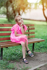 Little caucasian girl sitting in park on bench. A child in a beautiful pink dress in the open air