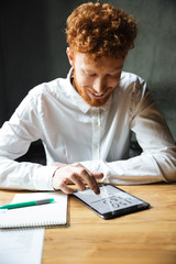 Close-up photo of young happy readhead bearded man using digital tablet at workplace