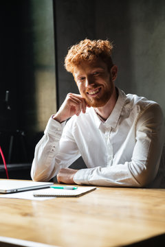 Young cheerful readhead bearded man in white shirt, sitting on his workplace, looking at camera