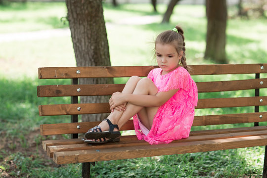 Little caucasian girl sitting in park on bench. A child in a beautiful pink dress in the open air - Powered by Adobe