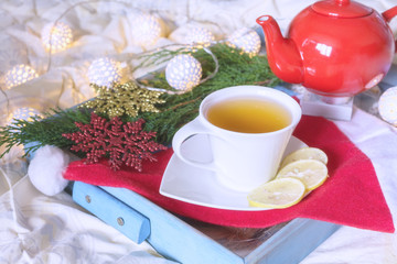 Breakfast with a cup of tea, a biscuit on a tray on the bed on a white blanket in the winter morning decorated with Christmas decorations, spruce branches with luminous garlands, the concept  holiday