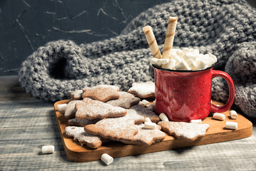 Red cup and gingerbread cookies. Mug of hot chocolate with marshmallow. Fresh baked star and fir-tree shaped biscuits on wooden background. Grey plaid or scarf