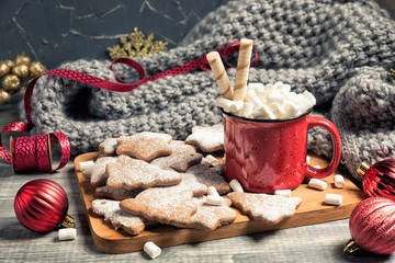 Red cup and gingerbread cookies. Mug of hot chocolate with marshmallow. Fresh baked star and fir-tree shaped biscuits on wooden background. Grey plaid or scarf