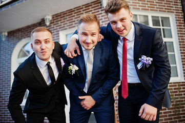 Handsome groom in his wedding tuxedo posing with groomsmen or best men outdoor with brick house on the background.