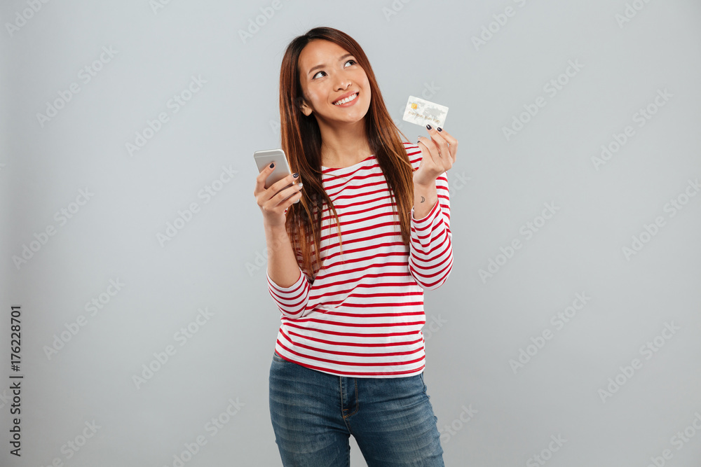 Poster Smiling pensive asian woman holding smartphone and credit card