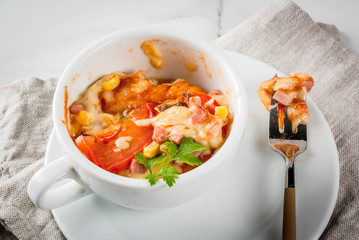 Trendy modern food, fast food, lunch in the microwave. Pizza in a mug, with ham, tomatoes, mushrooms, cheese. White cup, with a fork, on a white marble table. Copy space