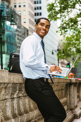 Young Happy African American Businessman traveling in New York, wearing white shirt, tie, black pants, carrying leather bag, working on laptop computer on street in Manhattan, looking up, smiling..