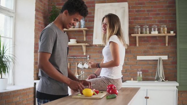 Handsome african guy is cutting vegetables preparing food while his caucasian girlfriend is sitting on the table in the kitchen in the morning. Happy multiethnic couple talking
