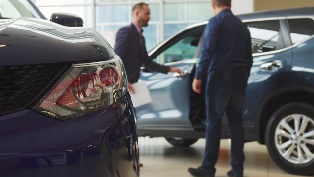 The seller leads the buyer to his new car. He opens the door to him. The buyer gets into the car in order to do a test drive