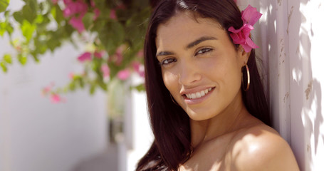 Obraz na płótnie Canvas Portrait of wonderful ethnic brunette smiling at camera while posing with fresh pink flowers in garden on background of sunny street.