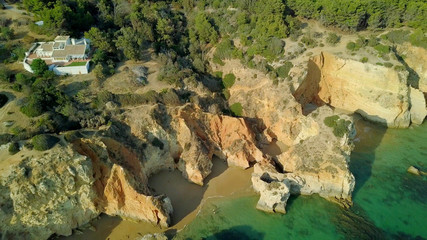 Aerial shot of colorful rocky cliffs and turquoise water of ocean washing sandy beach with mansion on hill above, Portugal, Algarve.