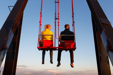 Young couple on the swing on the roof of tower at sunset time