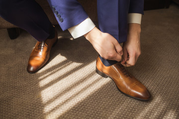 Young styish handsome man puts new brown modern comfortable leather shoes on in home sunny interior. Closeup view of male hands lacing beautiful elegant shoes. Horizontal color photography. - Powered by Adobe