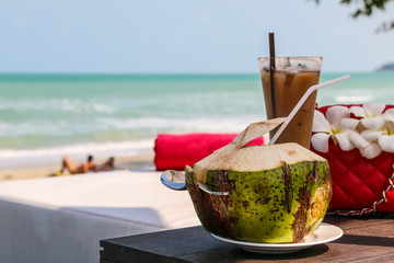 Coconut with drinking  straw, spoon  and cool coffee in the glass, red bag , beautiful white Plumeria flower on wooden table with blurred two people are sunbathing on the beach , blue sea and blue sky