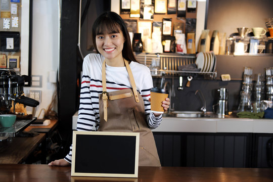 Smiling asian barista holding cup of coffee at counter in coffee shop. Cafe restaurant service, food and drink industry concept.